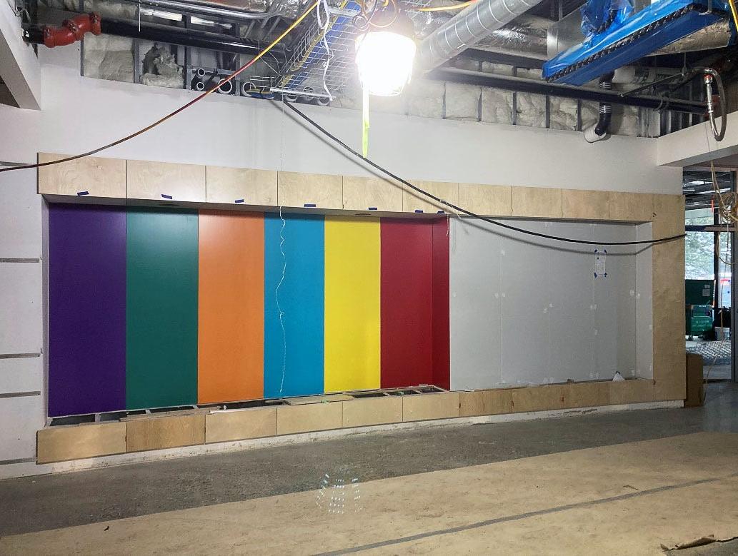a wall in a room under construction has colorful pieces of wod partially installed with light wood framing around the whole space