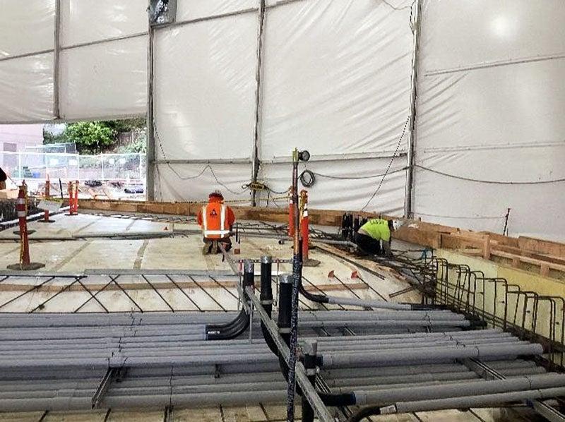 under a large tent, white waterproofing material has a stack of electrical conduit placed in a guie frame with rebar beneath it