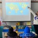 Two student teachers stand before elementary age students at a map of the world