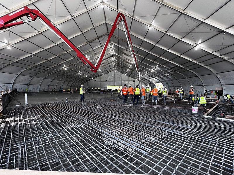 a grid of rebar is at the bottom, a group of workers in hardhats and safety vests stands in the center, a concrete pump is in the back with a large tent structure over it all