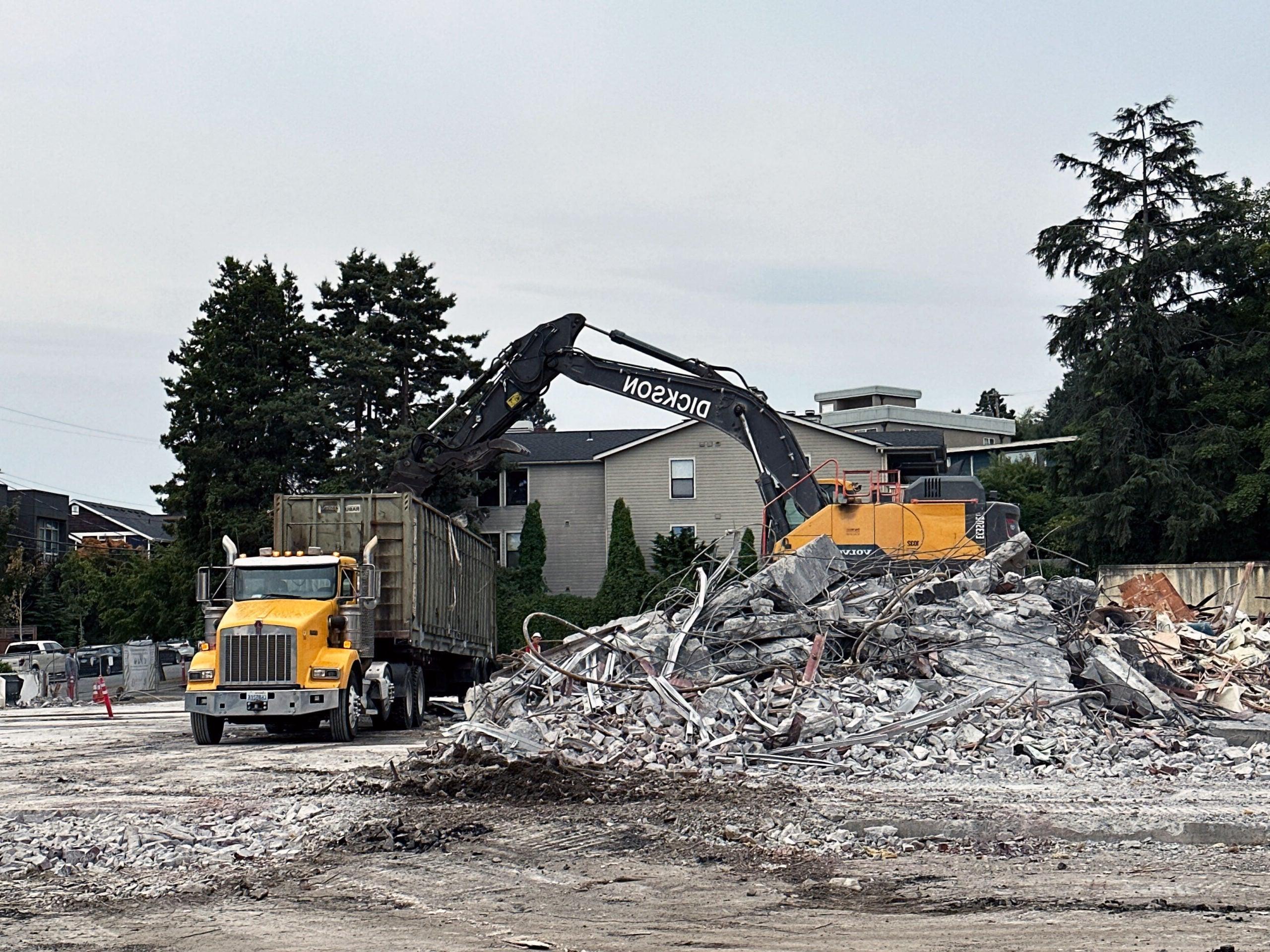 an excavator is on top of a pile of concrete rubble emptying into a container on a truck