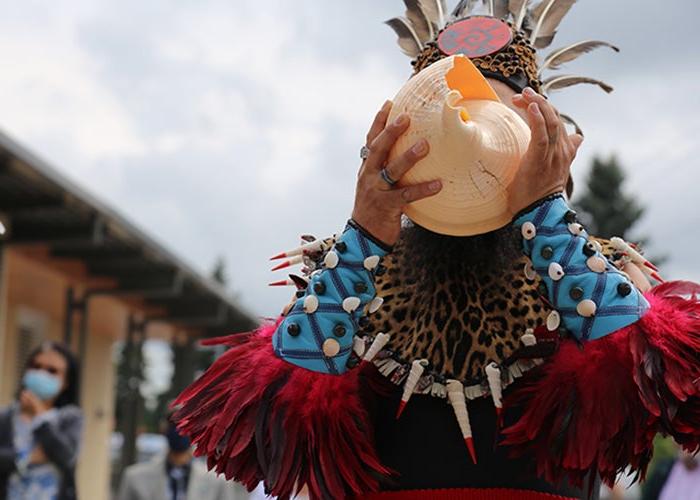 a person with a feather headdress, beaded cuffs, and a necklace with teeth holds a large pink shell in front of their face