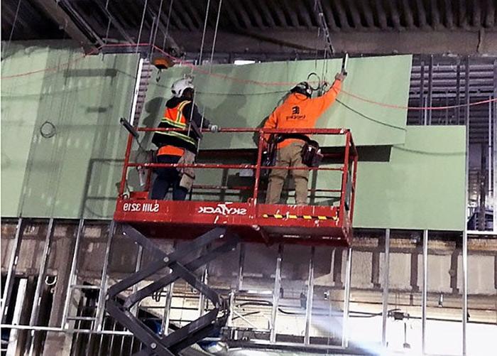 two workers in construction safety equipment are on a lift holding green sheets of material against studs